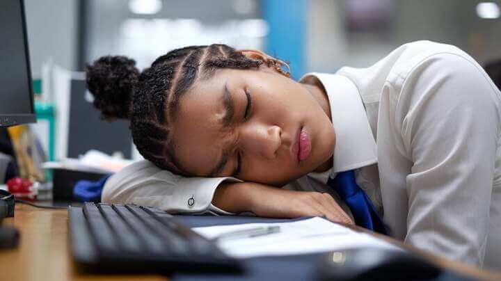 A young person resting their head on a desk in an office, looking tired and sleepy.
